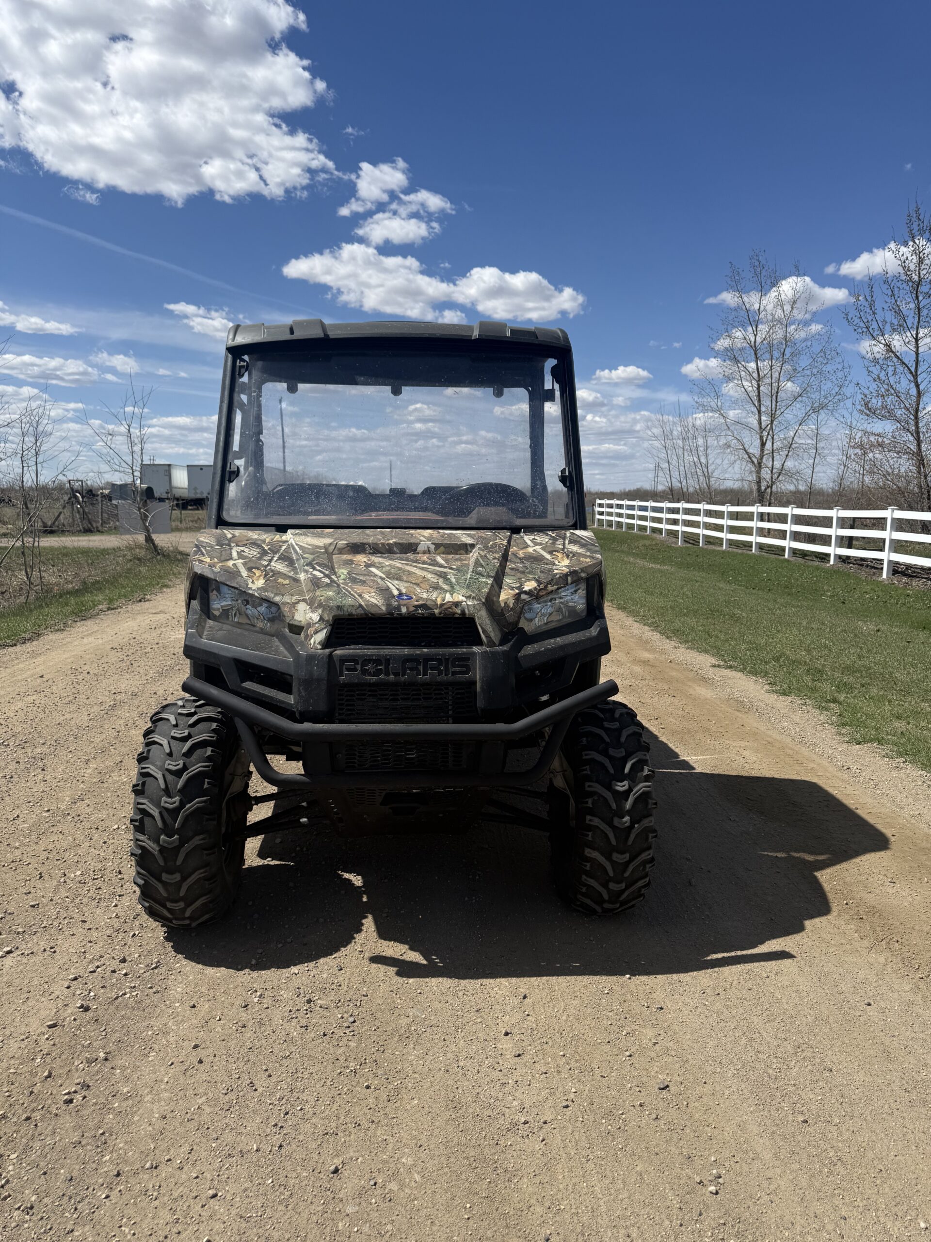 Front view of a camouflage Polaris off-road vehicle parked on a dirt road under a blue sky with scattered clouds.