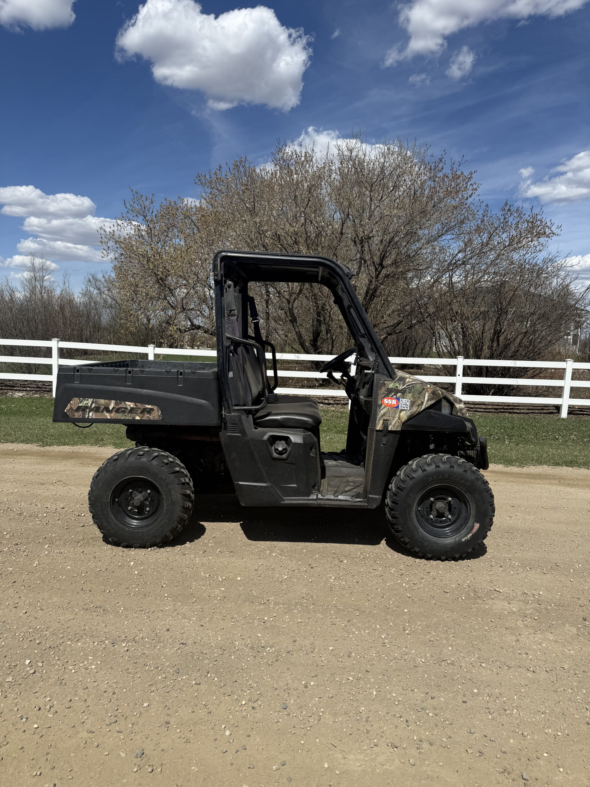 Black off-road utility vehicle (Ranger) parked on a dirt lot with leafless trees and a white fence under a blue sky misted with clouds.