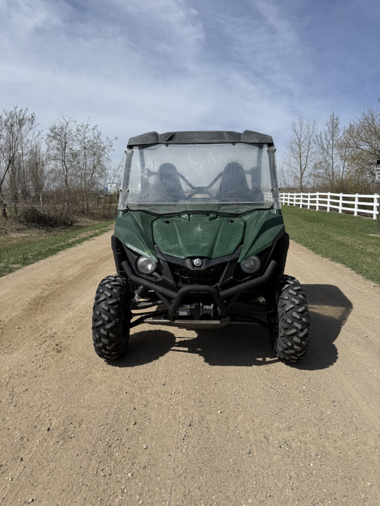Front view of a green off-road utility vehicle on a dirt road with a blue sky and bare trees in the background.