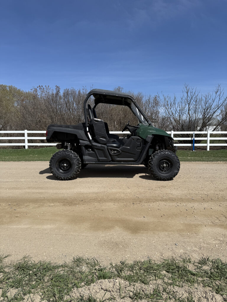 Green off-road utility vehicle parked on a dirt road with a white fence and bare trees under a clear blue sky.