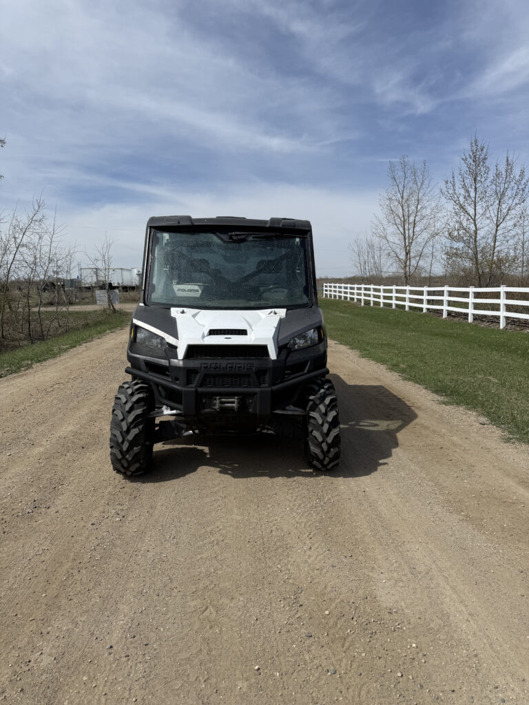 Front view of a black Polaris off-road utility vehicle on a dirt road, with a white fence and bare trees under a blue sky in the background.