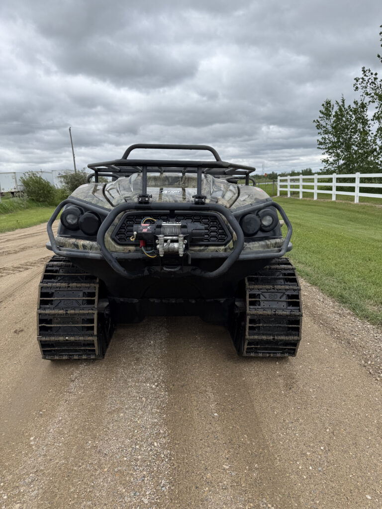 Front view of a tracked off-road vehicle with a heavy bumper and winch, parked on a dirt road; cloudy sky and rural fencing in background.