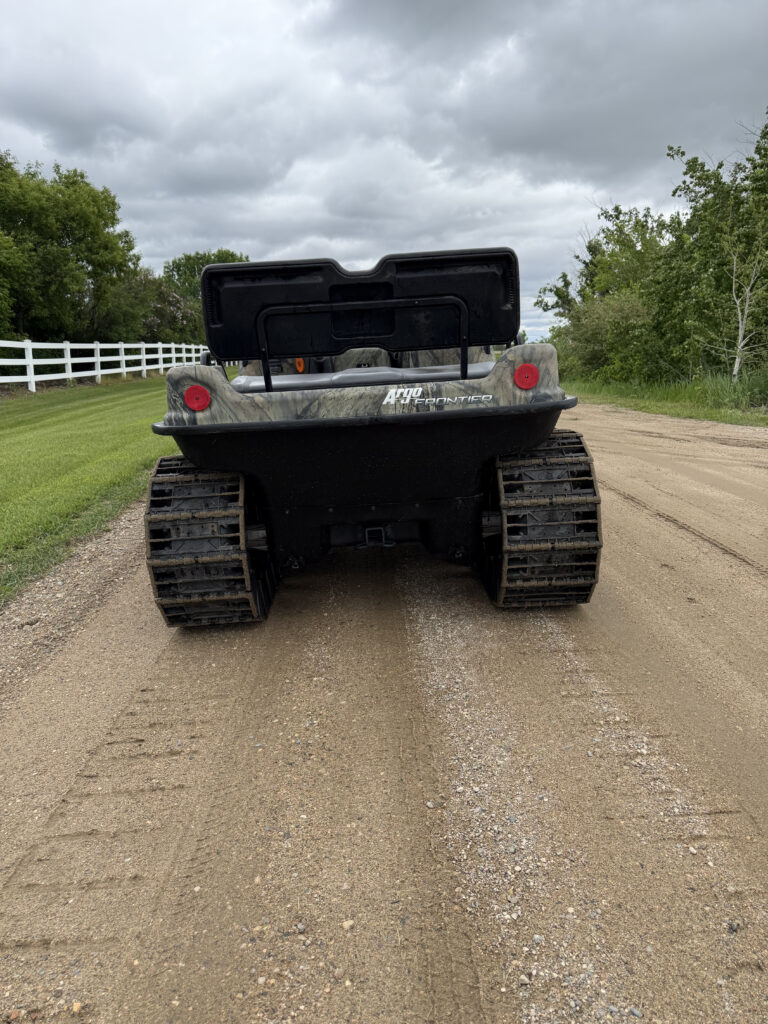 Rear view of an Argo Frontier all-terrain vehicle on a gravel road, with green fields and trees beside a white fence under a cloudy sky