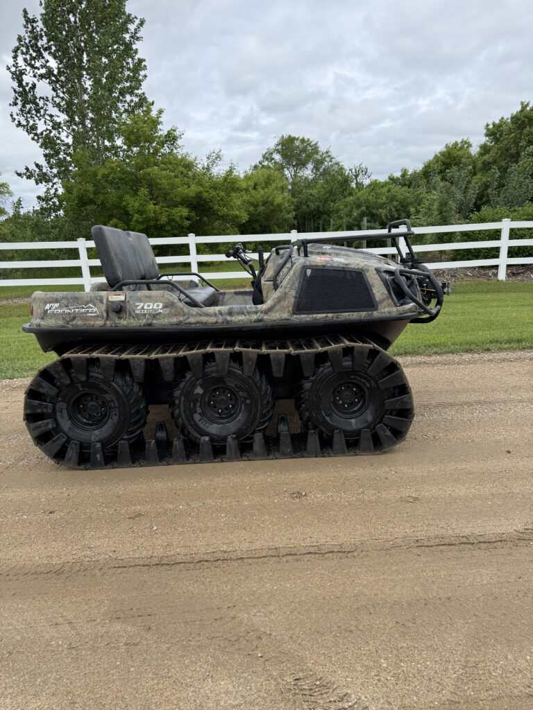 Camouflage Argo all-terrain vehicle with large track wheels parked on a dirt road, white fence and green trees in the background.